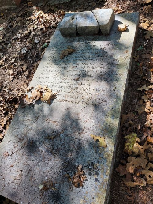 A weathered gravestone partially covered with leaves, inscribed with names and dates, set in a wooded area.