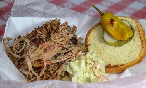 A plate featuring pulled pork, a sandwich bun with pickles and peppers, and a side of coleslaw on a checkered tablecloth.