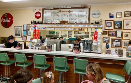 A vintage diner interior with a counter, green stools, and patrons enjoying food, featuring retro decor and a menu board.