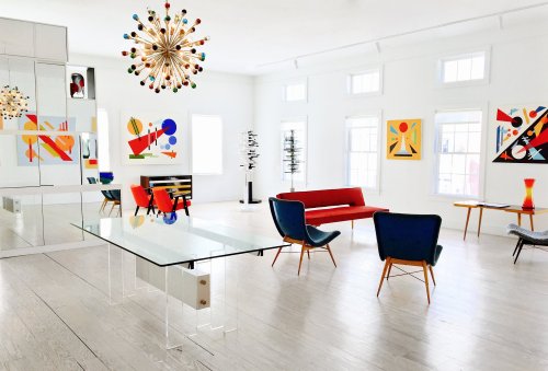 Bright, modern living room with colorful artwork, a glass table, red sofa, and stylish chairs against a white backdrop.