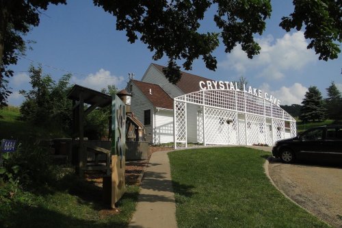 A white building with "CRYSTAL LAKE CAVE" sign, surrounded by greenery and a pathway leading to it.