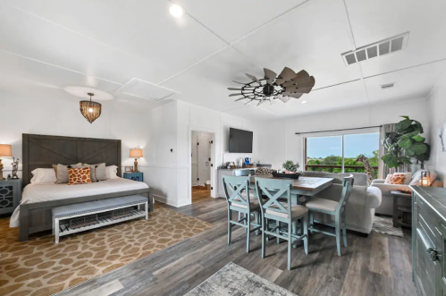 Cozy studio with a bed, dining area, and large window showcasing greenery outside. Modern decor and warm lighting.