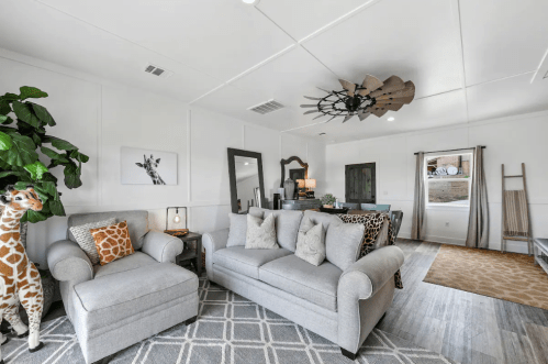 Cozy living room with gray furniture, giraffe decor, and a large ceiling fan, featuring a modern, inviting atmosphere.