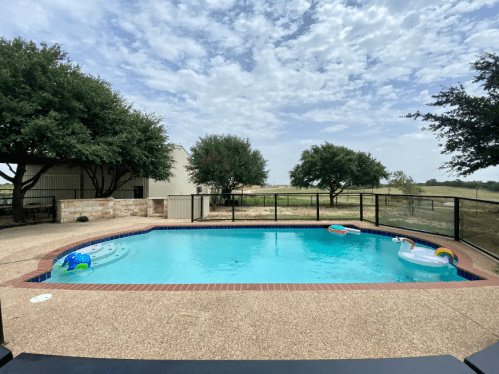 A clear blue pool surrounded by trees and a fence, with colorful floaties and a cloudy sky in the background.
