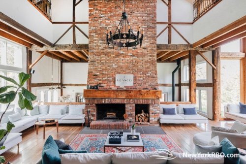 Spacious living room with a large brick fireplace, wooden beams, and modern furniture, featuring natural light and a cozy atmosphere.
