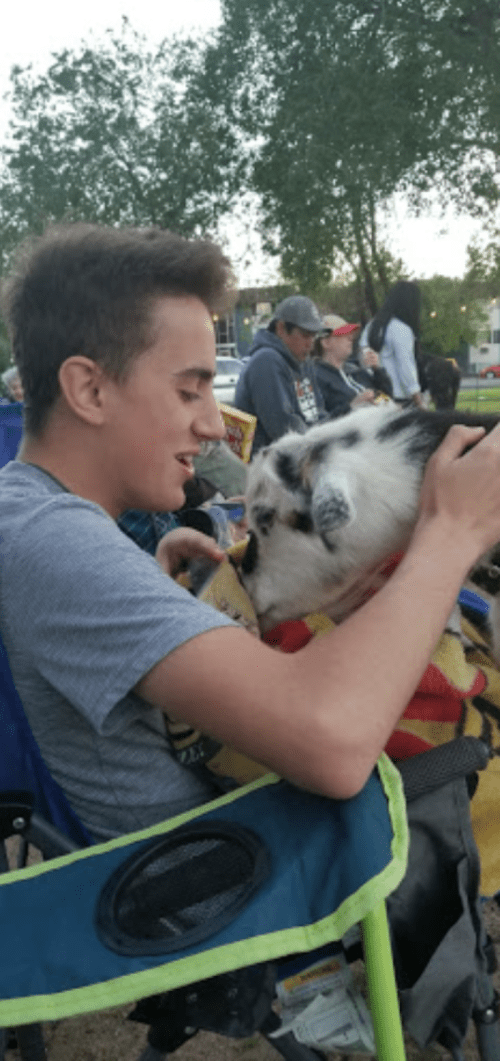 A young man sits in a chair, smiling and interacting with a dog wrapped in a colorful blanket.