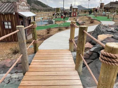 A wooden bridge leads to a mini-golf course with themed structures and people playing in the background.