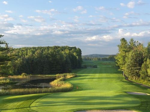 A scenic golf course with lush green fairways, trees, and a pond under a partly cloudy sky.