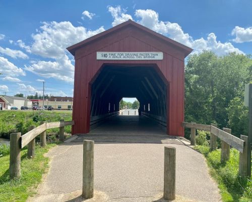 A red covered bridge with a pathway leading through it, surrounded by green trees and a blue sky with clouds.