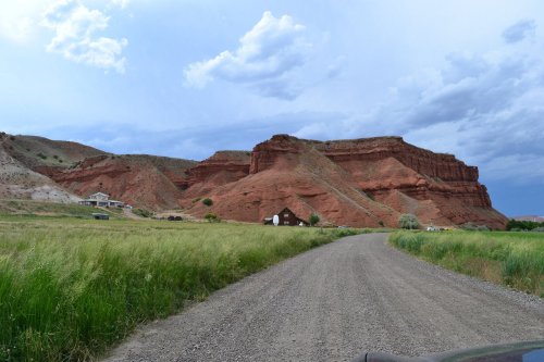 A gravel road leads through green grass to red rock formations under a cloudy sky. A small house is visible in the distance.