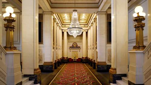 Elegant hallway with marble columns, a large chandelier, and a decorative carpet leading to a seating area.