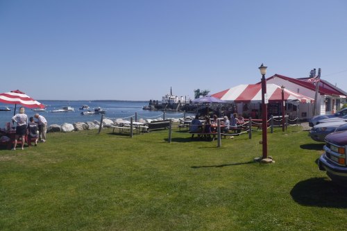A sunny waterfront scene with boats, a striped canopy, and people dining at picnic tables by the shore.
