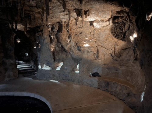 A dimly lit cave interior with textured rock walls, illuminated by soft lights, and a curved stone bench.