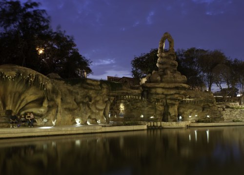 A serene nighttime scene of a rocky structure by a calm water body, surrounded by trees and softly lit pathways.