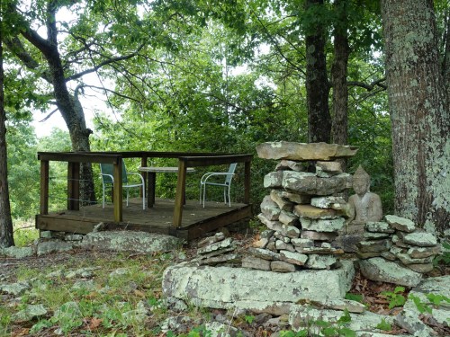 A serene outdoor space with a wooden deck, two chairs, and a stone Buddha statue surrounded by trees and rocks.