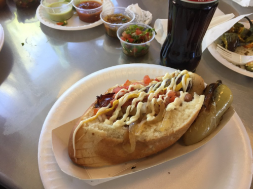 A hot dog topped with onions, tomatoes, and sauces, served with a side of jalapeños and a soda.