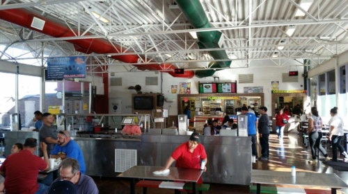 A busy food court with people dining, staff cleaning tables, and colorful signage in a bright, open space.