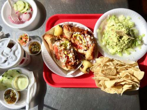 A red tray with two loaded hot dogs, guacamole, lettuce, tortilla chips, and side dishes of cucumbers and radishes.