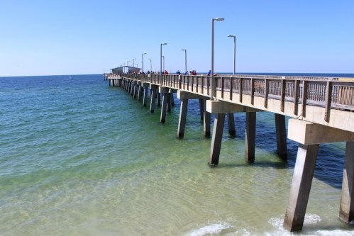 A wooden pier extends over calm blue waters, with people walking along it under a clear sky.
