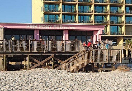 A beachside pub with a pink exterior and wooden deck, surrounded by sand and a tall hotel in the background.