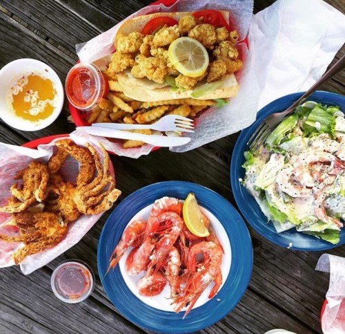 A spread of seafood dishes including shrimp, a fish sandwich, fries, and a salad on a wooden table.