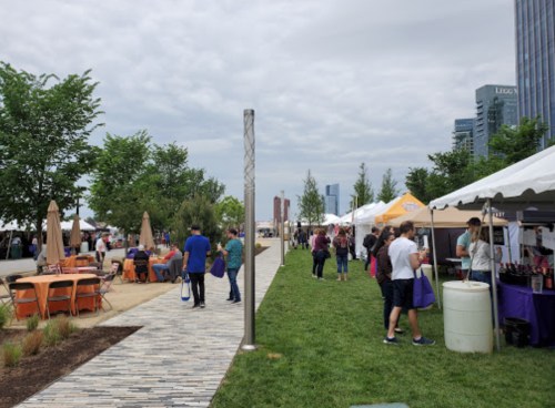 A bustling outdoor market with tents, people walking, and greenery under a cloudy sky.