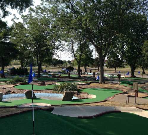 A mini-golf course with green turf, trees, and blue flags, featuring various holes and water features.