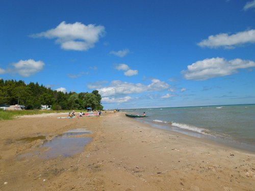 A sandy beach with a calm lake, scattered clouds, and people relaxing in the distance under a blue sky.
