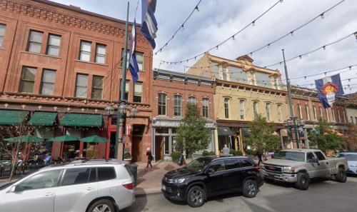 A street view of historic buildings with flags, string lights, and parked cars in a lively urban area.