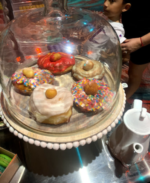A glass dome displays colorful donuts with sprinkles and toppings on a wooden platter, with a child in the background.