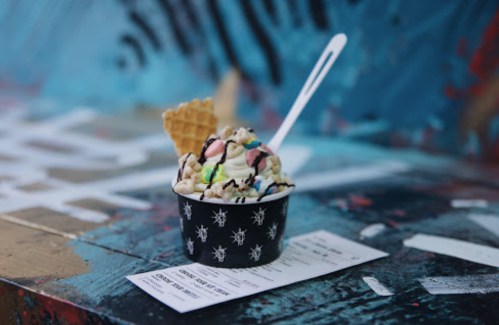 A cup of colorful ice cream topped with whipped cream, chocolate drizzle, and a waffle cone, resting on a ticket.