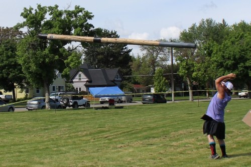 A person in a purple tank top throws a heavy pole in a grassy field, with trees and houses in the background.