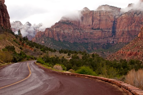 A winding road through a lush valley, surrounded by towering red rock cliffs and misty clouds in the background.
