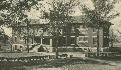 Historic brick building with a porch, surrounded by trees and a manicured lawn. Black and white photo.