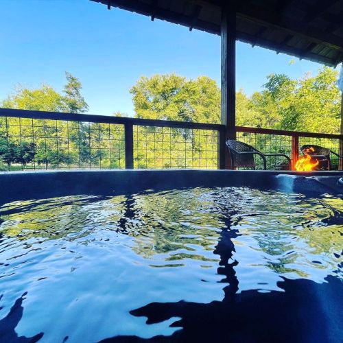 A serene outdoor scene featuring a hot tub with a view of trees and a fire pit under a clear blue sky.