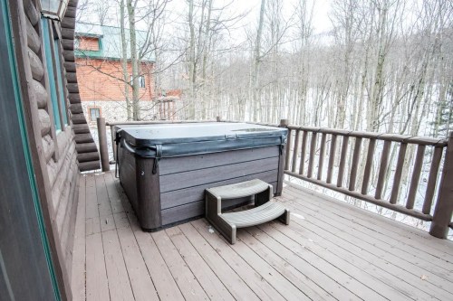 A wooden deck with a hot tub and steps, surrounded by trees in a snowy landscape. A cabin is visible in the background.