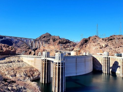 A large dam surrounded by rocky terrain and a clear blue sky, with a bridge in the background.