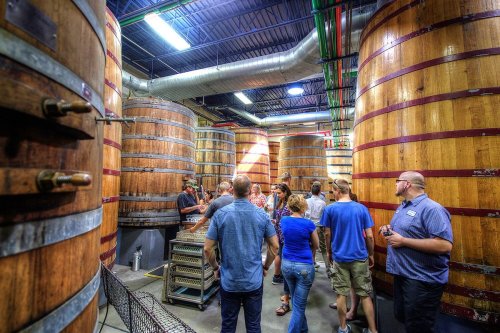 A group of people tours a brewery, surrounded by large wooden fermentation tanks.