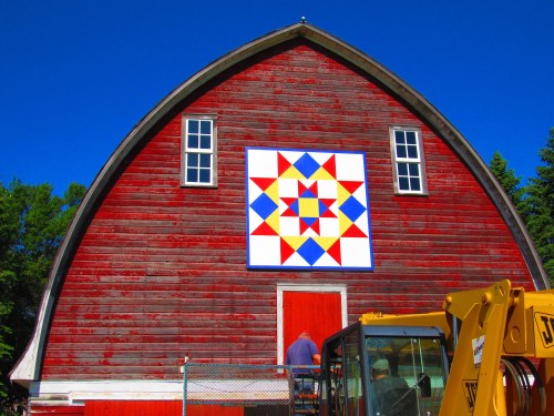 A red barn with a colorful quilt pattern on its side, under a clear blue sky. Two people are near the barn.