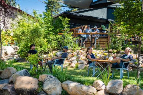 A serene outdoor scene with people sitting at tables among greenery and rocks, enjoying a sunny day.