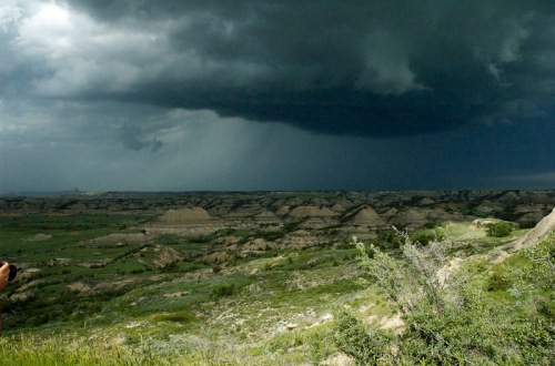 Dark storm clouds loom over a rugged landscape with rolling hills and green vegetation.