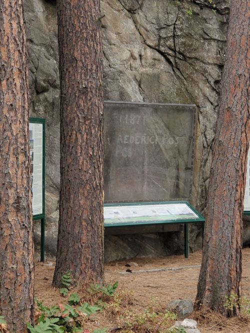Information display near a rocky surface, surrounded by tall trees, featuring text about Frederick Post from 1871.