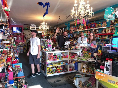 A colorful toy store filled with shelves of toys, three people smiling at the camera, and chandeliers hanging above.
