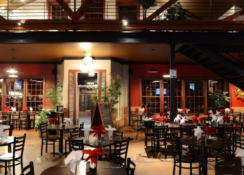 Cozy restaurant interior with wooden tables, chairs, and festive decorations, featuring plants and a staircase in the background.
