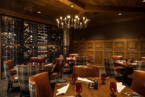 Cozy restaurant interior with wooden accents, elegant lighting, and a wine cellar in the background. Red glasses on tables.