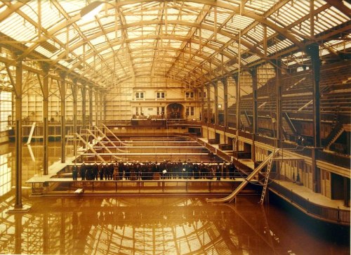 Historic indoor swimming pool with a large glass roof, featuring spectators gathered on a balcony above the water.
