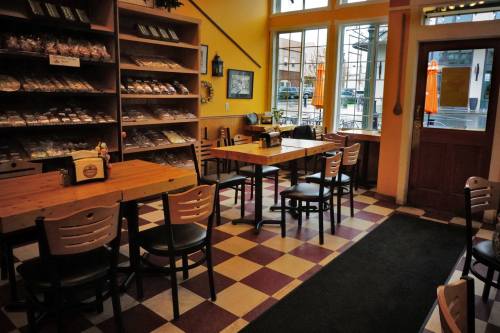 Cozy café interior with wooden tables, chairs, and shelves filled with treats, featuring large windows and a welcoming atmosphere.