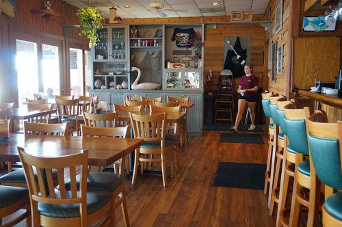 A cozy restaurant interior with wooden tables, chairs, and a server walking by, featuring nautical decor and a welcoming atmosphere.