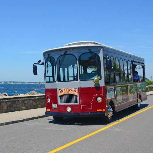 A colorful trolley bus driving along a scenic coastal road with a clear blue sky and water in the background.