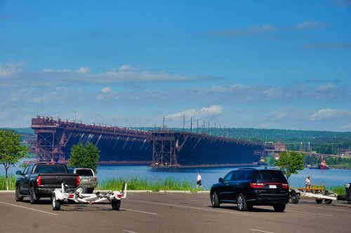 A view of a large industrial dock over water, with parked vehicles in the foreground and a clear blue sky.
